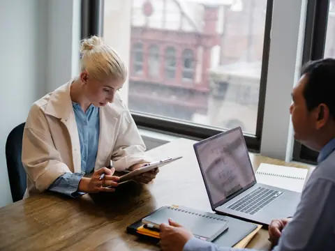 A woman reading papers at a wooden desk with a laptop in a bright office
