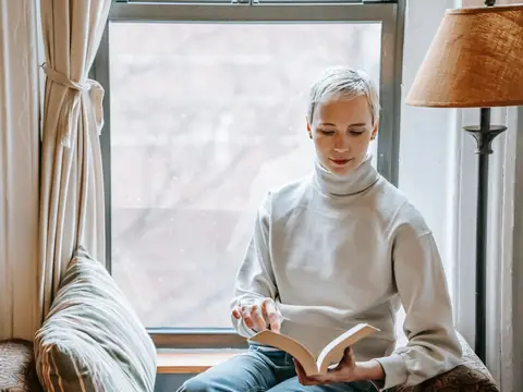 A woman reading a book on a cushioned seat by a bright window