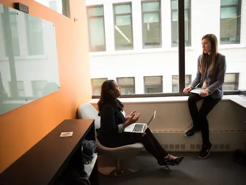 Two women talking by a row of bright windows, one with a laptop