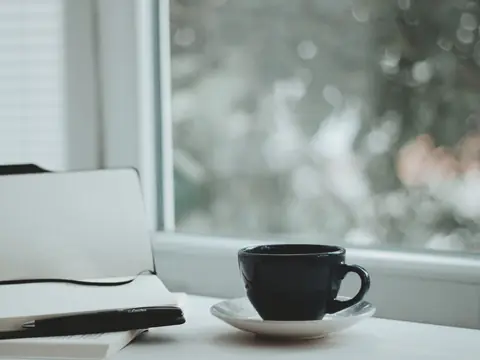 A notebook, a pen and a cup of coffee on a white table by a window