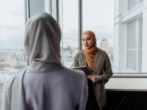 Two women talking by a tall window with a city view, one holding a tablet