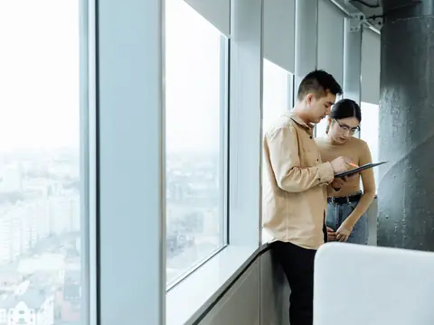 Two people looking over a tablet beside a tall window above the city