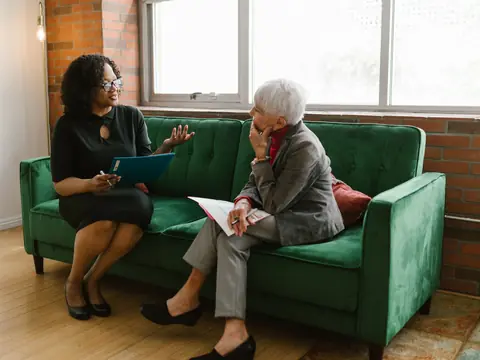 Two women talking over papers on a green sofa in a bright room